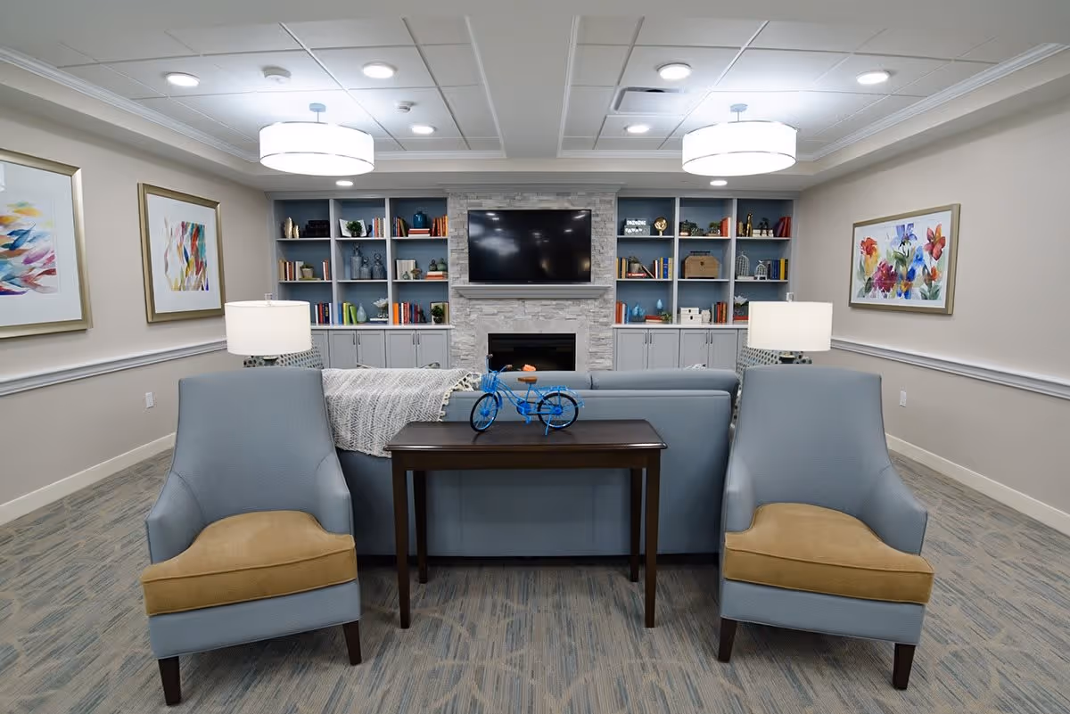 A cozy living room with two light blue armchairs with tan cushions facing a blue sofa. Behind the sofa is a dark wooden console table with a small blue decorative bicycle on it. The far wall features built-in blue shelves filled with books and decorative items, a stone fireplace, and a mounted flat-screen TV. The room is softly lit by ceiling lights and two table lamps on either side of the sofa. Colorful abstract paintings hang on the beige walls.