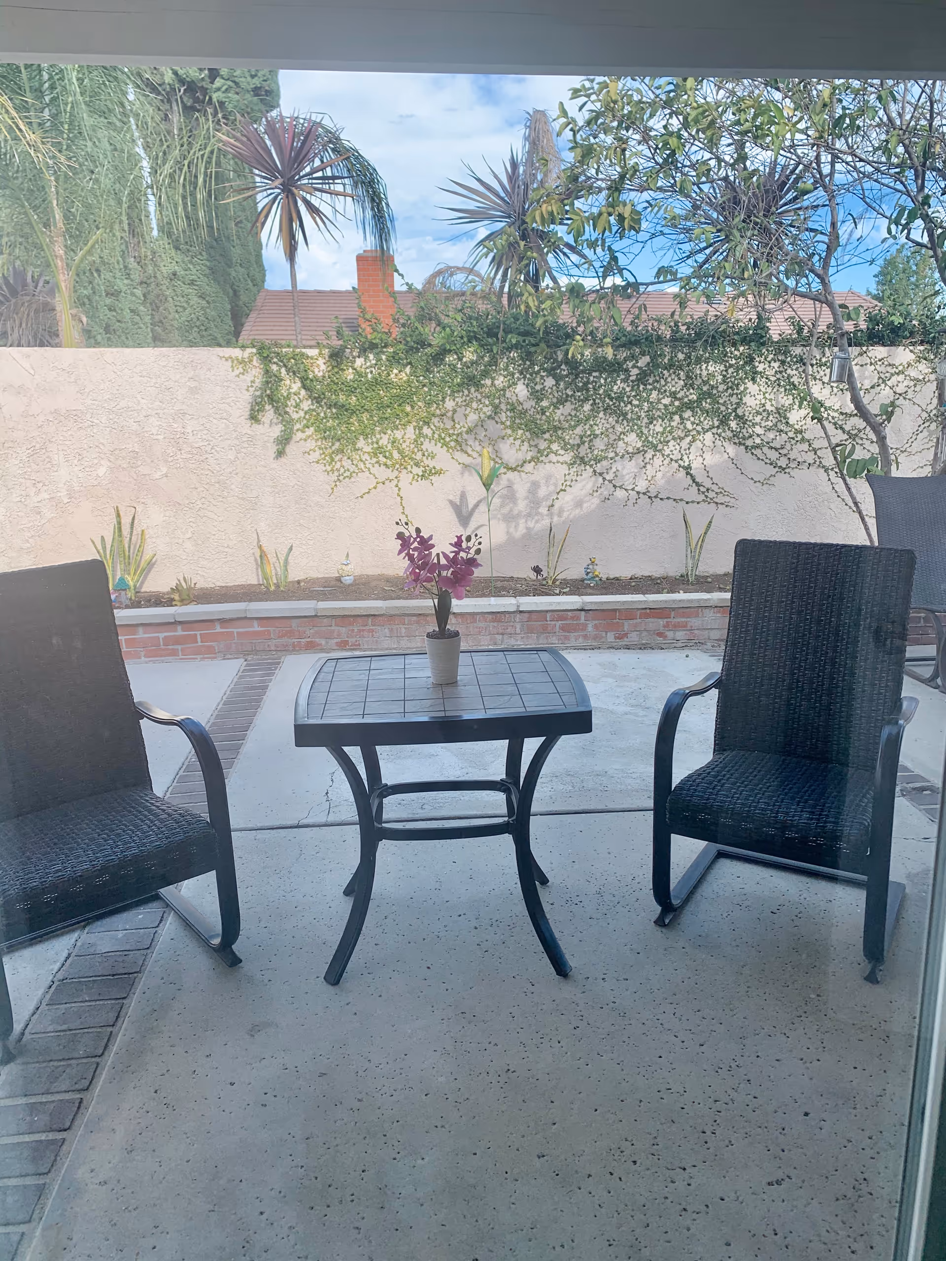 Outdoor patio area with two black wicker chairs and a small square table with a potted plant with purple flowers on top. The patio is surrounded by a low brick wall and a beige stucco wall with greenery and trees visible beyond it under a partly cloudy sky.