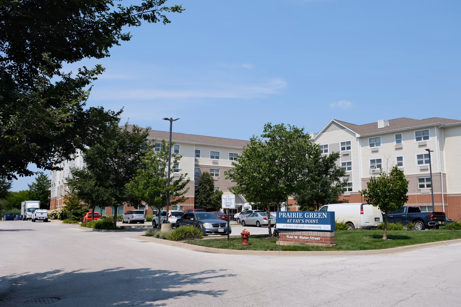 Exterior view of a senior living community named Prairie Green at Fay's Point, showing a multi-story building with beige and brick facade, surrounded by trees and a parking lot with several cars parked. A blue sign in front displays the community name and address 1546 W. Water Street.