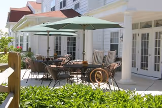 Outdoor patio area at a senior living facility with tables, chairs, and large green umbrellas. The patio is adjacent to a white building with multiple glass doors and windows. There are green bushes in the foreground and rocking chairs near the building.