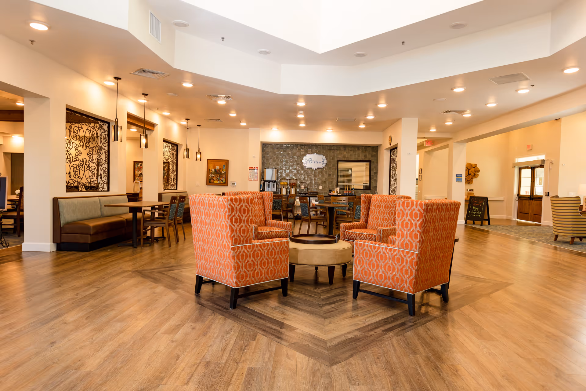 A spacious and well-lit common area with four orange patterned armchairs arranged around a small round table in the center. The room features wood flooring with a decorative inlay, several dining tables and chairs along the walls, pendant lights hanging from the ceiling, and a beverage station labeled 'Bistro' at the back. The space has a warm and inviting atmosphere with light-colored walls and decorative wall art.