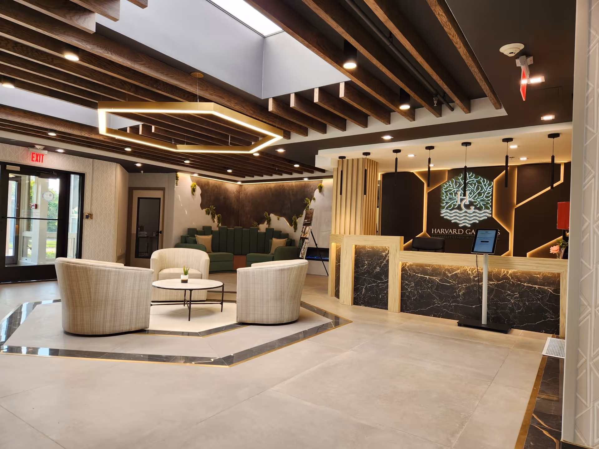 Modern and spacious reception area of Harvard Gardens Rehabilitation and Care Center featuring a marble and wood front desk with illuminated logo on the wall, a seating area with four beige armchairs around a small round table, green cushioned bench seating along the back wall, wooden ceiling beams, and large glass doors leading outside.
