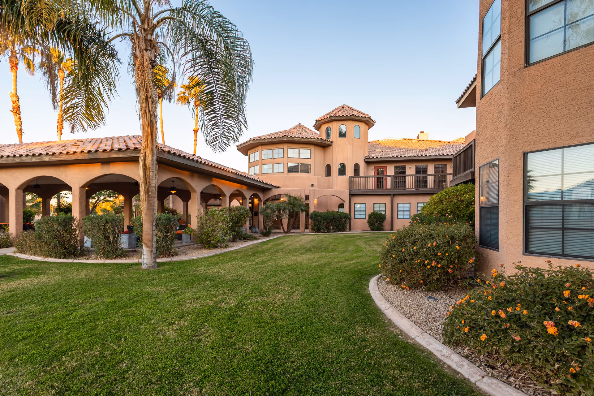 Exterior view of a senior living facility with a well-maintained green lawn, palm trees, and bushes with orange flowers. The building features a stucco finish with arched walkways, multiple windows, and a tiled roof under a clear sky.
