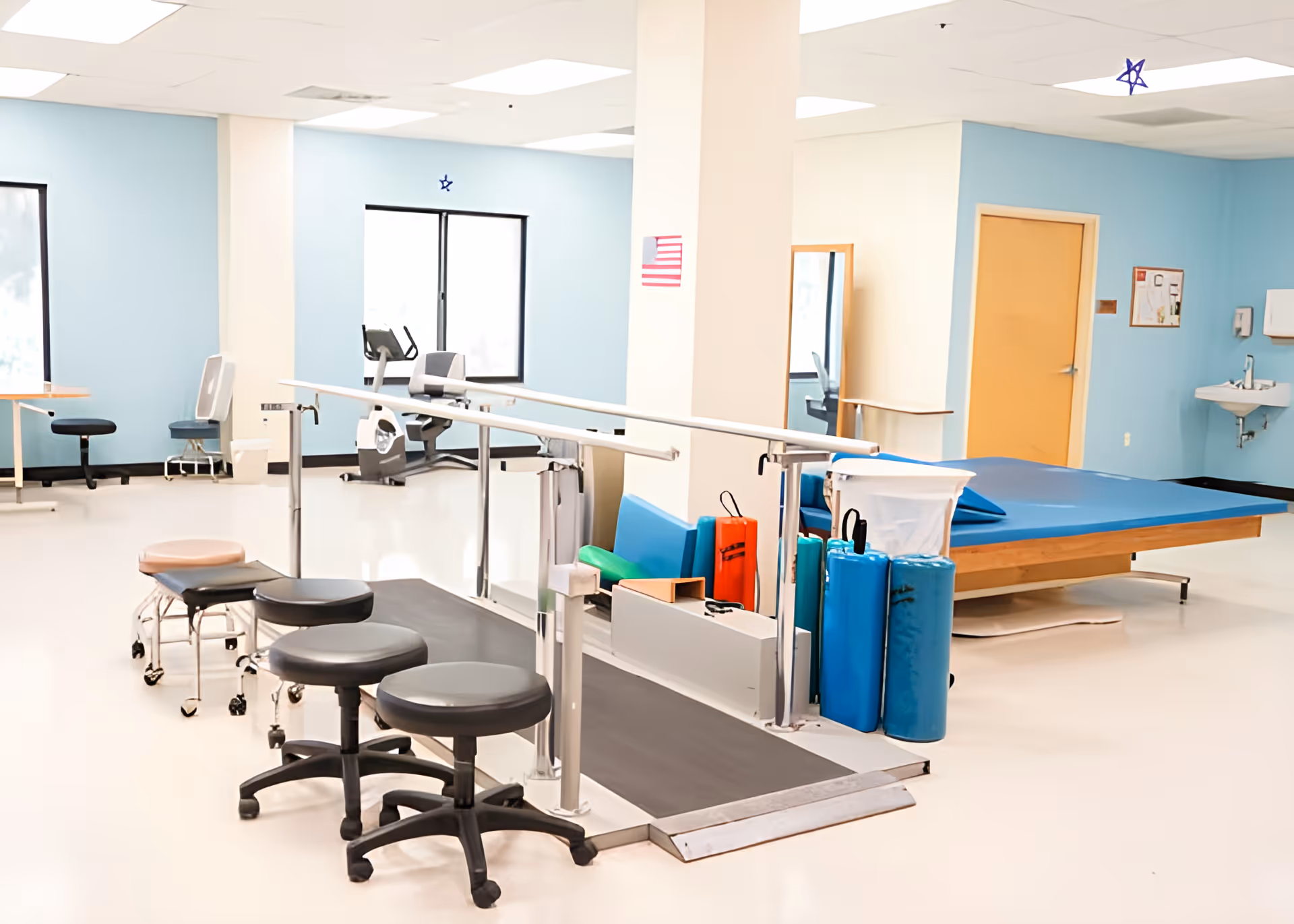 A bright rehabilitation room with light blue walls, featuring parallel bars for walking exercises, several rolling stools, a large padded therapy table, exercise equipment including stationary bikes, and a wall-mounted sink. The room has large windows allowing natural light to enter.