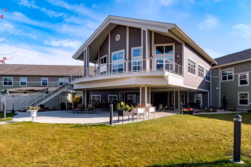 Exterior view of a two-story senior living facility building with a covered patio area on the ground floor and a balcony on the upper floor. The building has large windows and is surrounded by a grassy lawn with some outdoor seating and planters. The sky is clear and blue.