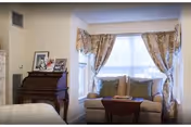 A cozy living area with a small sofa adorned with cushions in front of a window with floral curtains. To the left, there is a wooden roll-top desk with framed photos and decorative items on top. Part of a bed is visible in the foreground.