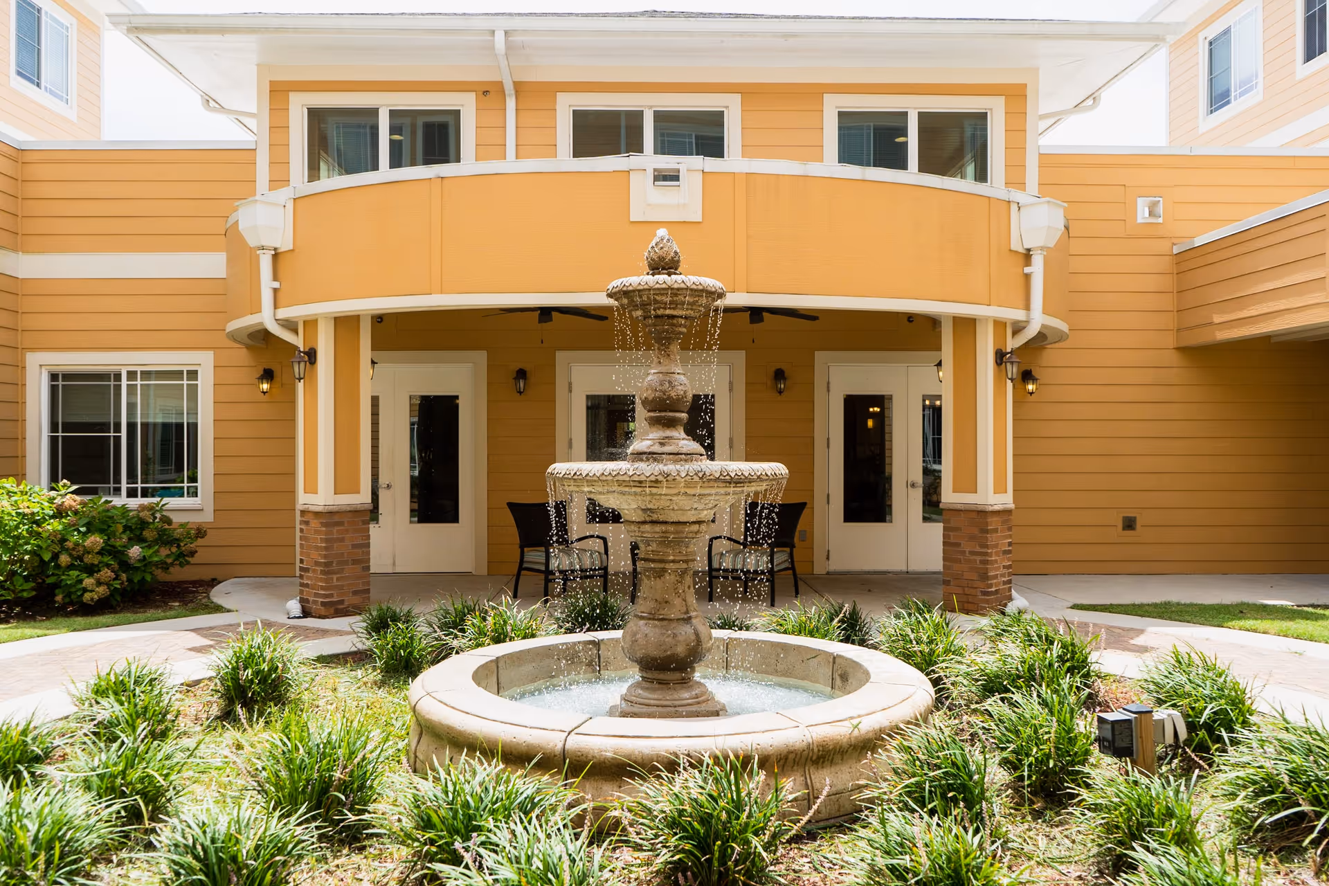 Outdoor courtyard area of a senior living facility with a multi-tiered stone water fountain in the center surrounded by green plants. The building has a yellow exterior with white trim, multiple windows, and two white doors leading inside. There are black chairs and tables under a covered patio area behind the fountain.