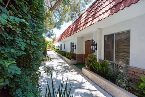 White single-story building with red-tiled roof and a sunlit walkway lined by greenery
