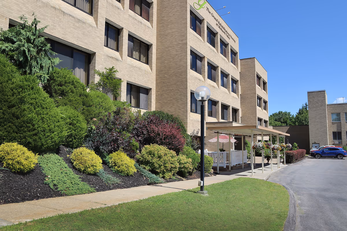 Front exterior of a multi-story brick senior rehabilitation and nursing facility with landscaped shrubs, a covered entrance porch, and parked cars.