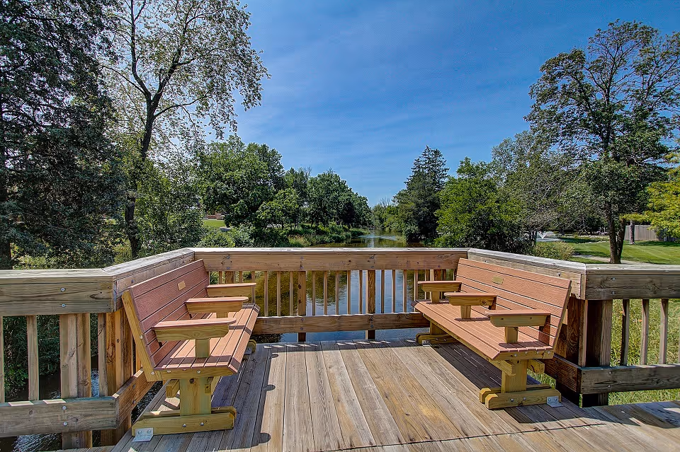 A wooden deck with two wooden benches facing each other overlooking a calm river surrounded by green trees under a clear blue sky.