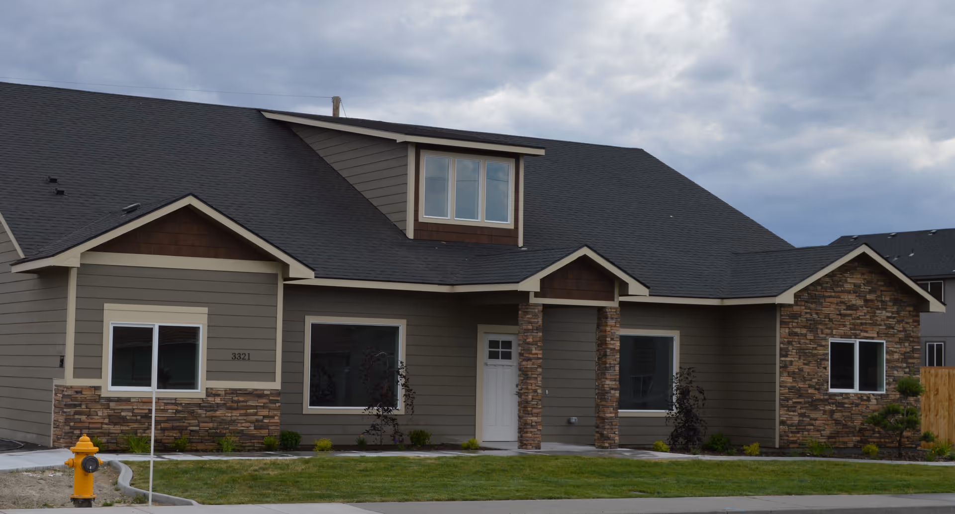 Front exterior view of a single-story residential building with gray siding, stone accents, a black roof, and a white front door. There is a green lawn and a yellow fire hydrant in front of the house under a cloudy sky.