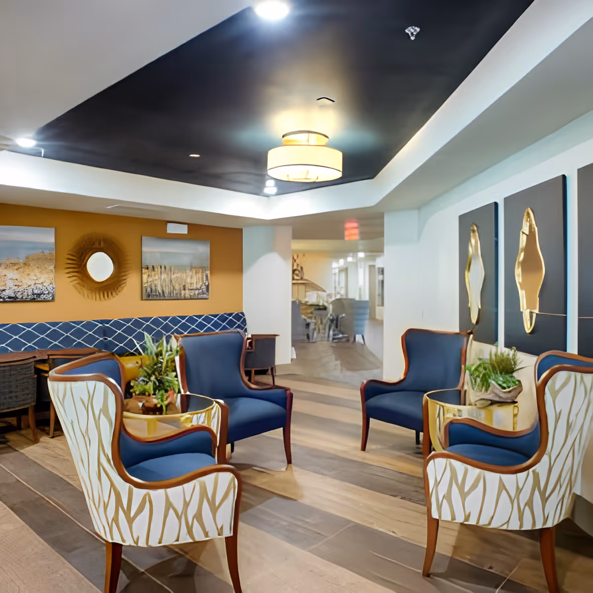 A modern and elegant seating area in a senior living facility featuring four armchairs with blue cushions and patterned white backs arranged around two small round tables with plants. The room has a dark ceiling with recessed lighting and a central hanging light fixture. The walls are decorated with abstract art and a circular mirror, and the floor has a wood-like finish.