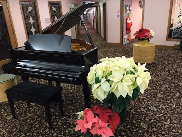 Interior view of a hallway in a senior living facility with a black grand piano and piano bench on a patterned carpet. There are white and red poinsettia plants placed near the piano and on a round wooden table. The hallway extends into the distance with doors and framed artwork on the walls.