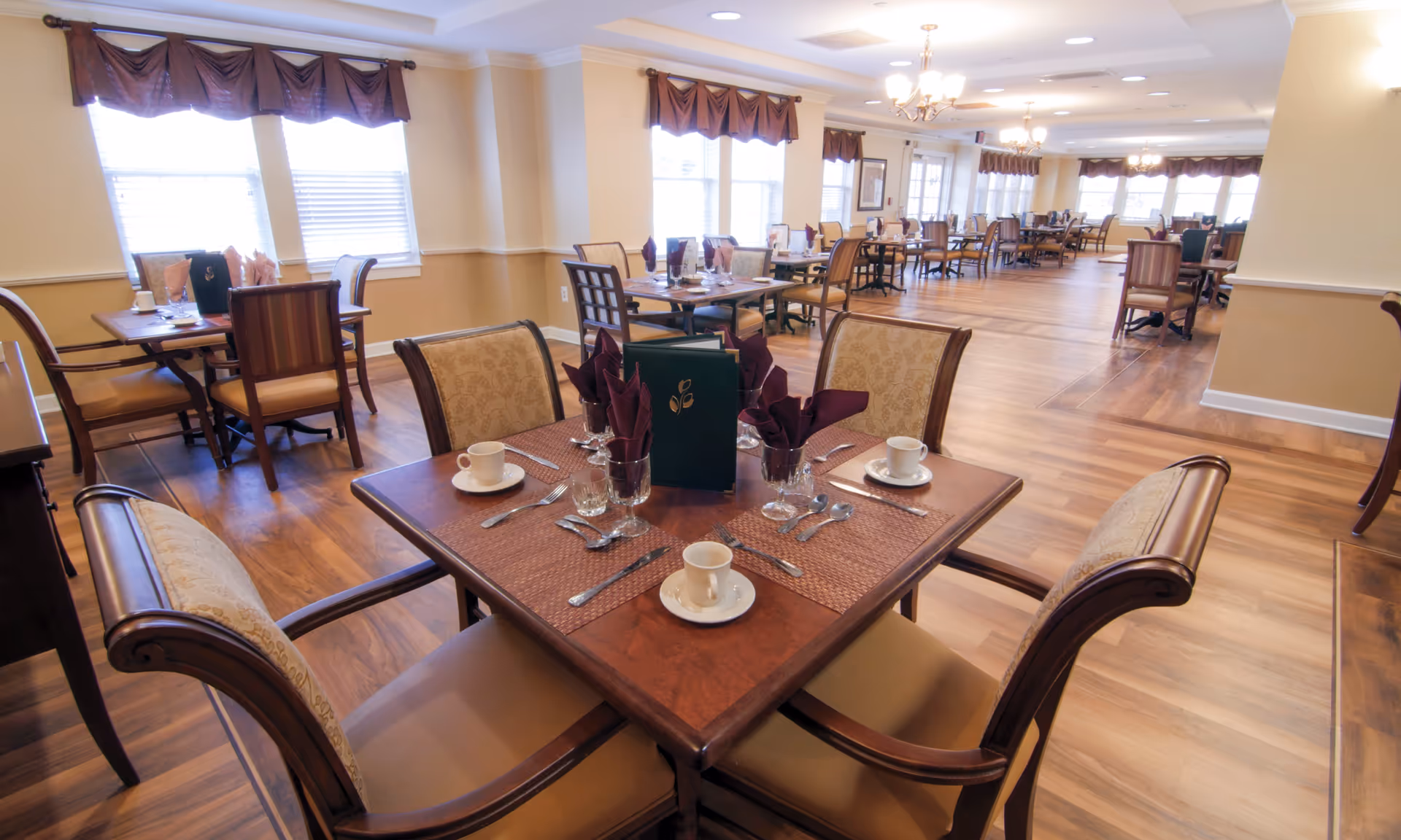 A spacious dining room with multiple wooden tables and cushioned chairs arranged neatly. Each table is set with cups, saucers, silverware, glasses, and folded napkins. Large windows with valances allow natural light to brighten the room, which has wood flooring and soft yellow walls.