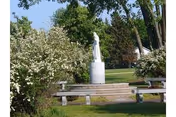 Outdoor garden area with a white statue on a pedestal surrounded by greenery and flowering bushes, with benches arranged around the statue.