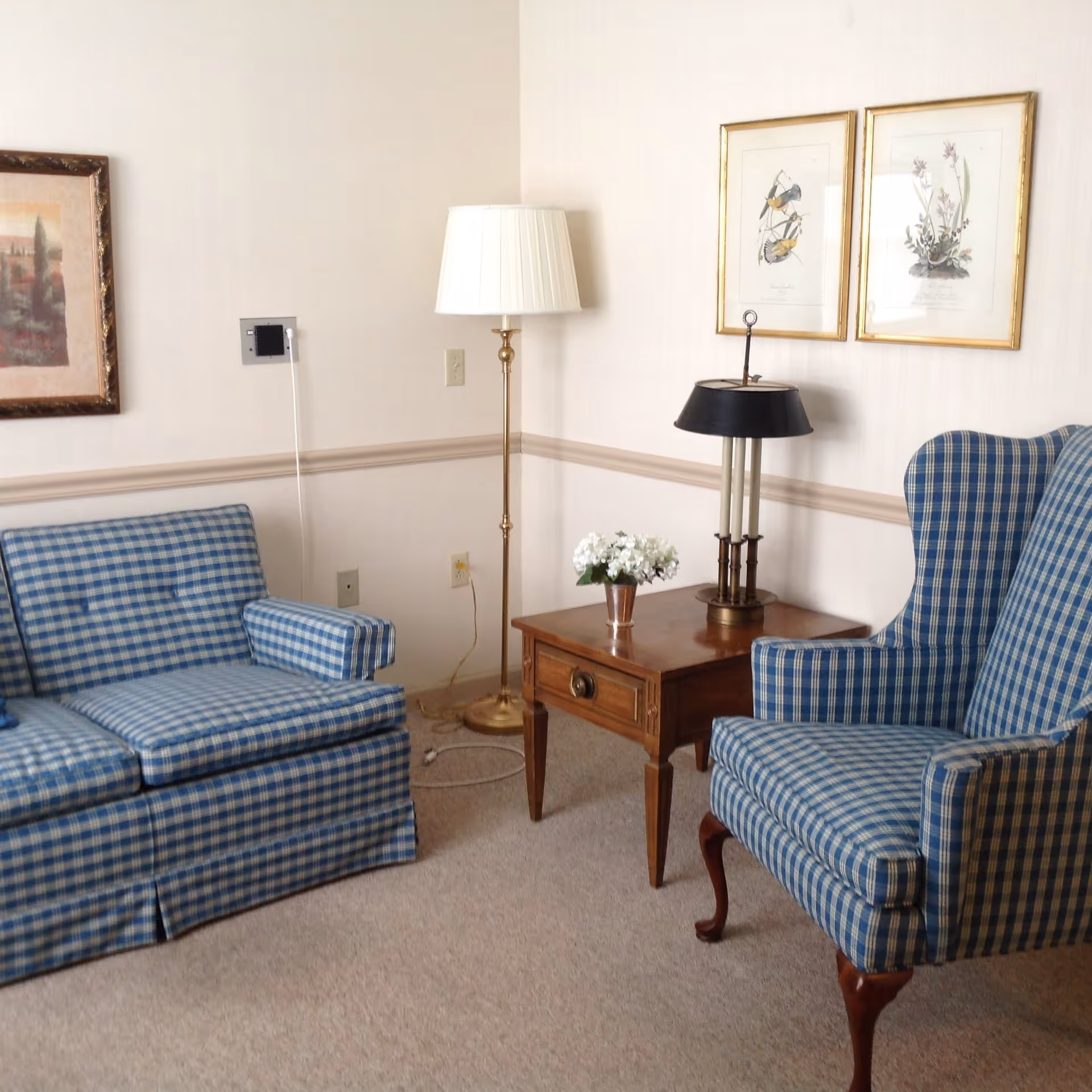 A cozy sitting area with a blue and white checkered loveseat and matching armchair. Between them is a wooden side table with a small flower arrangement and a lamp with a black shade. A tall floor lamp with a white shade stands behind the loveseat. Two framed botanical prints hang on the wall above the armchair, and another framed picture is on the adjacent wall.
