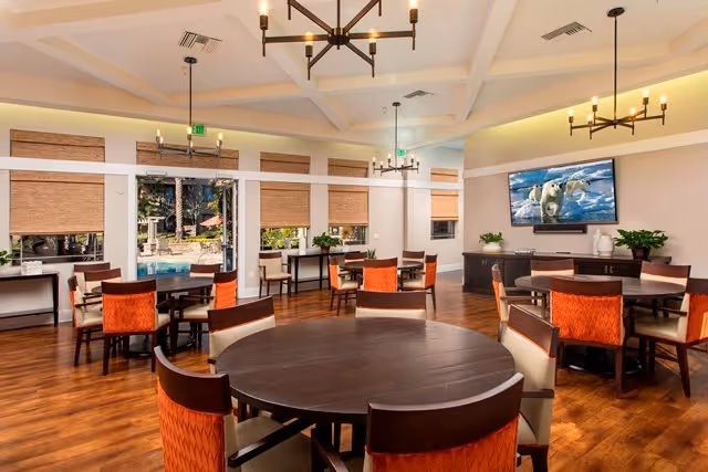 Bright communal dining room with round wooden tables, orange-upholstered chairs, chandeliers, large windows, and a wall-mounted TV.