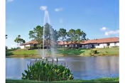 A scenic outdoor view of a pond with a water fountain in the center, surrounded by greenery and plants. In the background, there is a single-story building with a red-tiled roof and several trees under a blue sky with a few clouds.