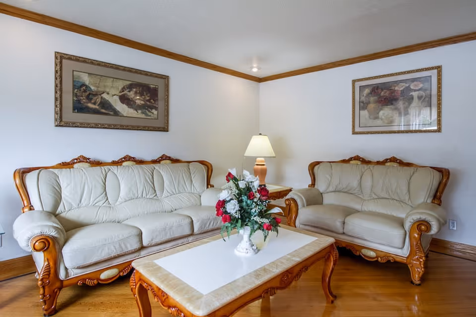 A living room with two cream upholstered sofas, a wooden coffee table with a floral centerpiece, a table lamp, and framed artwork on the walls.