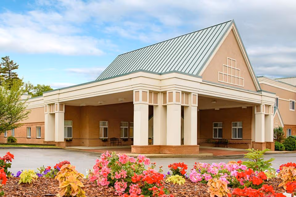 Front exterior view of a senior living facility building with a large covered entrance supported by columns. The building has a light brown facade with white trim and a green metal roof. In the foreground, there is a landscaped area with colorful flowers and plants under a partly cloudy sky.