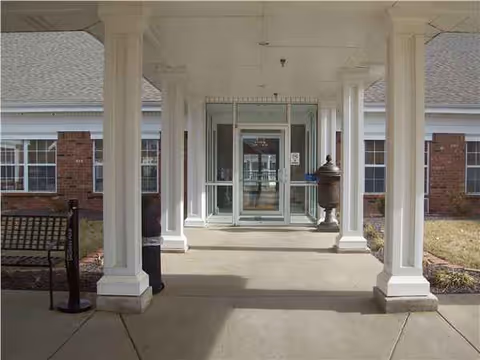 Columns line a covered walkway toward glass doors at the entrance of a senior living facility.