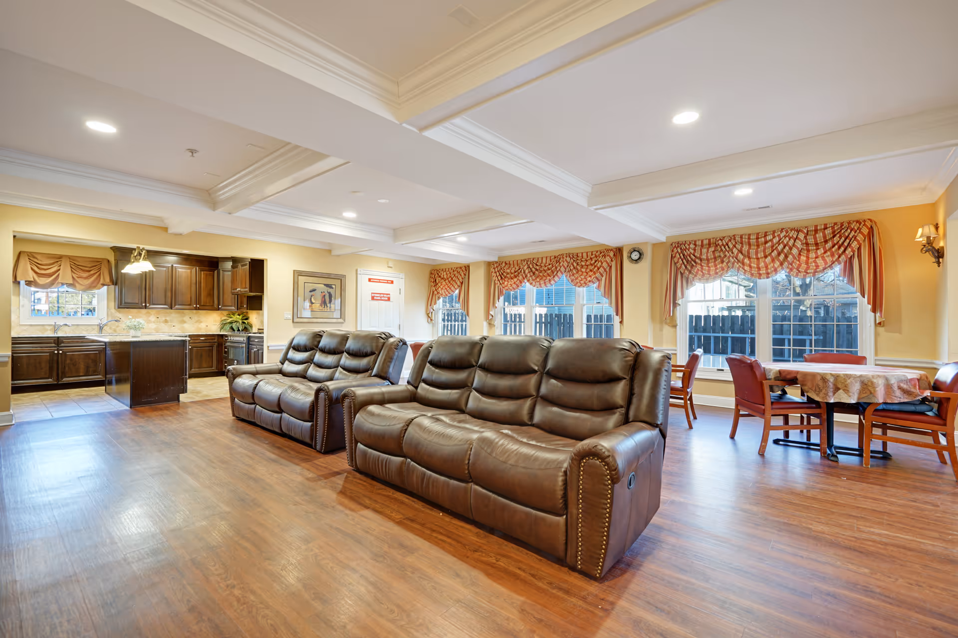 A spacious and well-lit common area featuring two brown leather sofas facing away from a kitchen area with dark wood cabinets. The room has large windows with patterned valances, wooden flooring, and a dining table with chairs near the windows. The ceiling has recessed lighting and decorative beams.