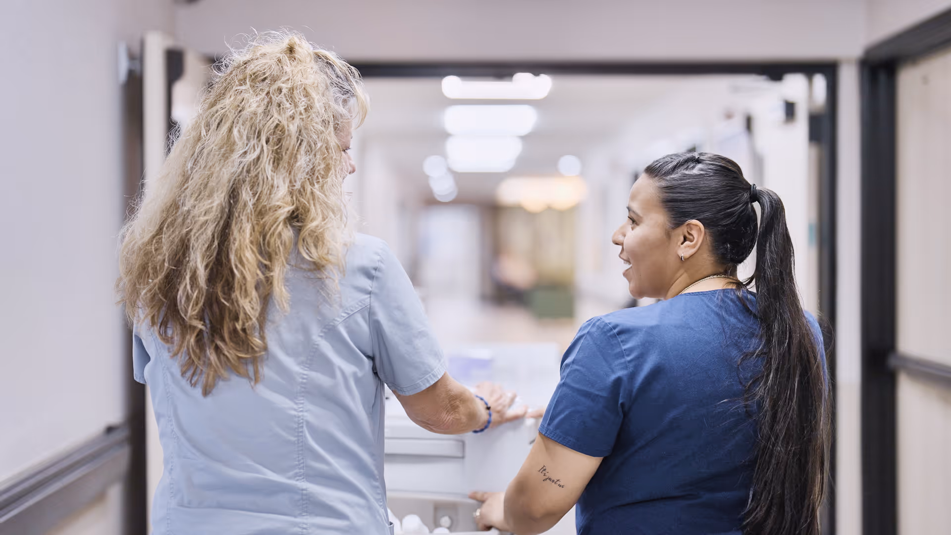 Two healthcare workers in scrubs walking down a hallway in a medical or rehabilitation facility, seen from behind, engaged in conversation while pushing a cart.