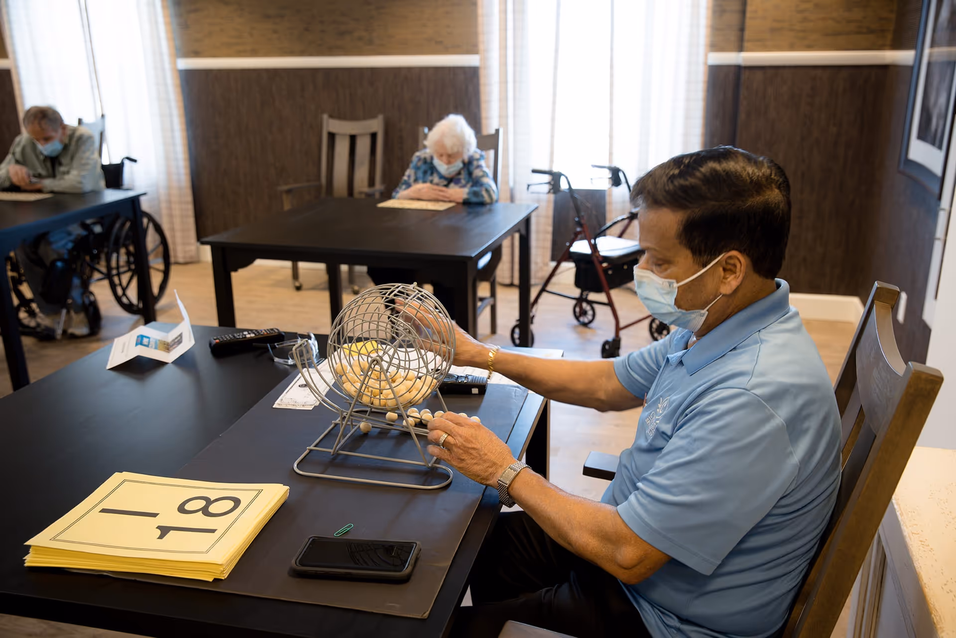 An elderly man wearing a mask is sitting at a table playing bingo with a bingo cage and balls in front of him. In the background, two other elderly individuals, also wearing masks, are seated at separate tables in a well-lit room with wooden chairs and light-colored curtains. A walker is visible near the window.