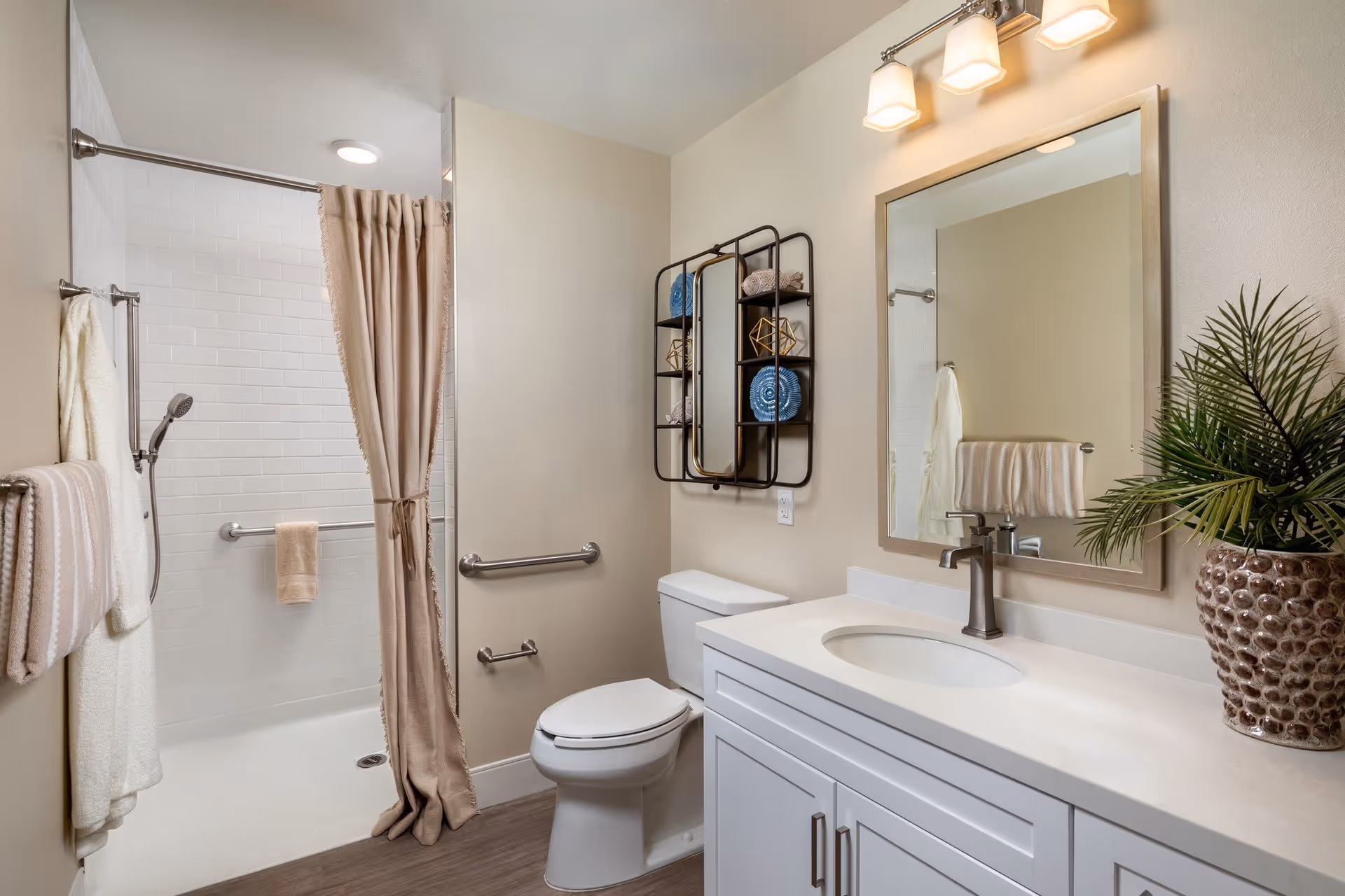 A clean and modern bathroom with a white vanity and sink, a large mirror above the sink, a toilet, and a shower area with a beige curtain. The shower has white subway tiles and a handheld showerhead. There are beige and white towels hanging on towel bars, a decorative wall shelf with blue and gold accents, and a potted plant on the vanity countertop.