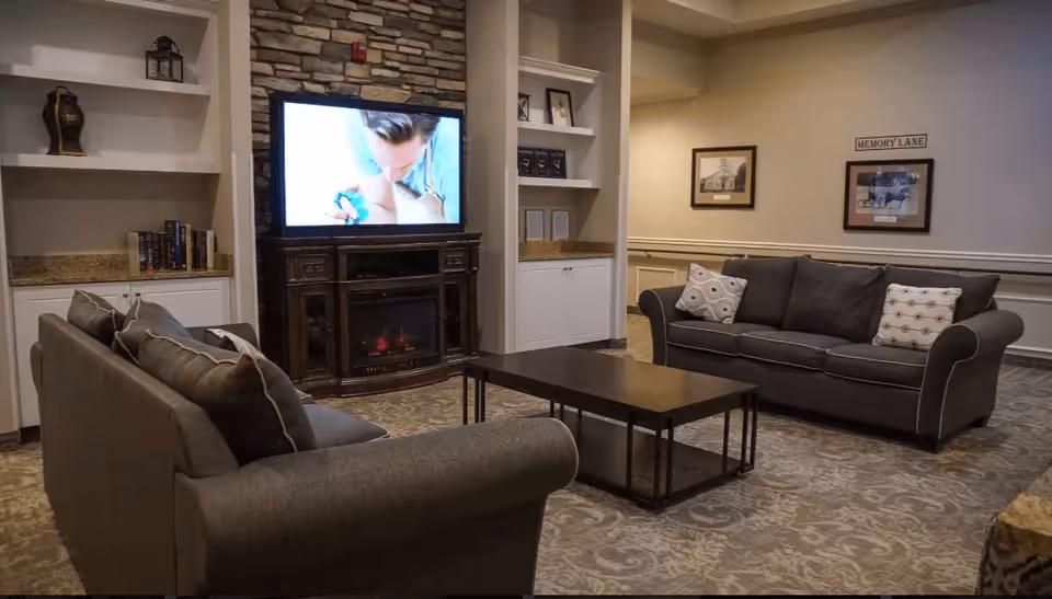 A cozy living room with two gray sofas facing each other, a dark wooden coffee table in the center, and a flat-screen TV mounted above a fireplace with a stone backdrop. Built-in shelves with decorative items and books flank the fireplace. The walls have framed pictures and a sign that reads 'MEMORY LANE'.