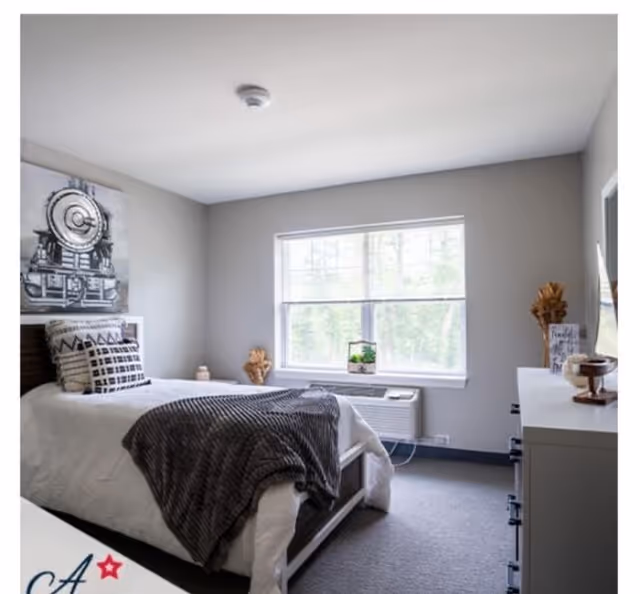 A neatly made single bed with white bedding and a dark gray throw blanket in a bright bedroom. The room has a large window letting in natural light, a dresser with decorative items, and a framed picture of a train on the wall.