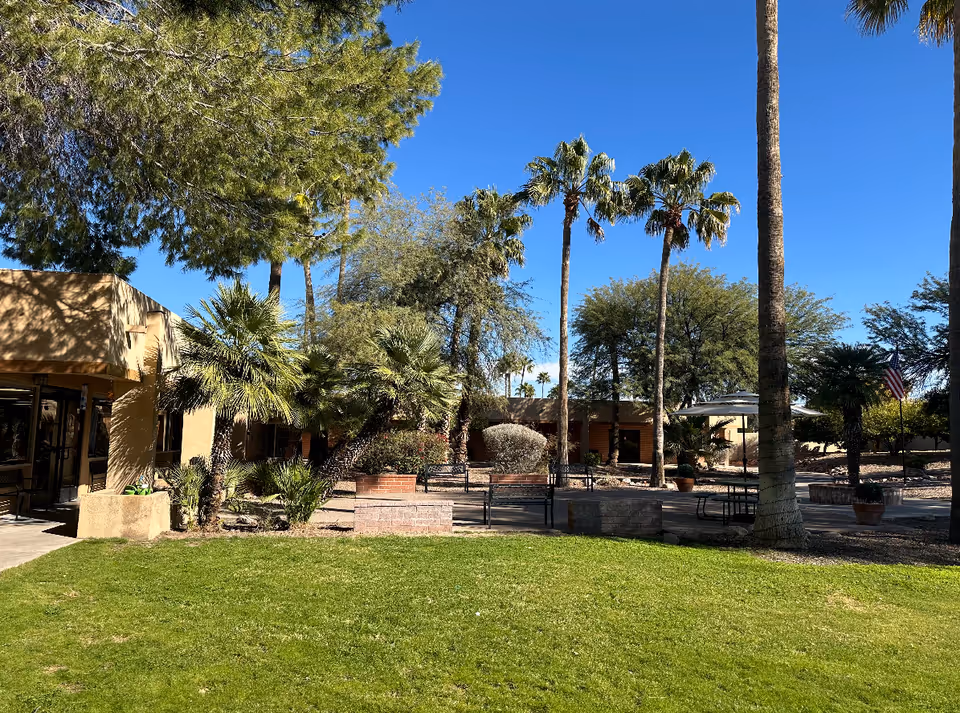 Outdoor garden area at Woodland Palms Memory Care featuring green grass, palm trees, other trees and shrubs, benches, a table with an umbrella, and a clear blue sky.
