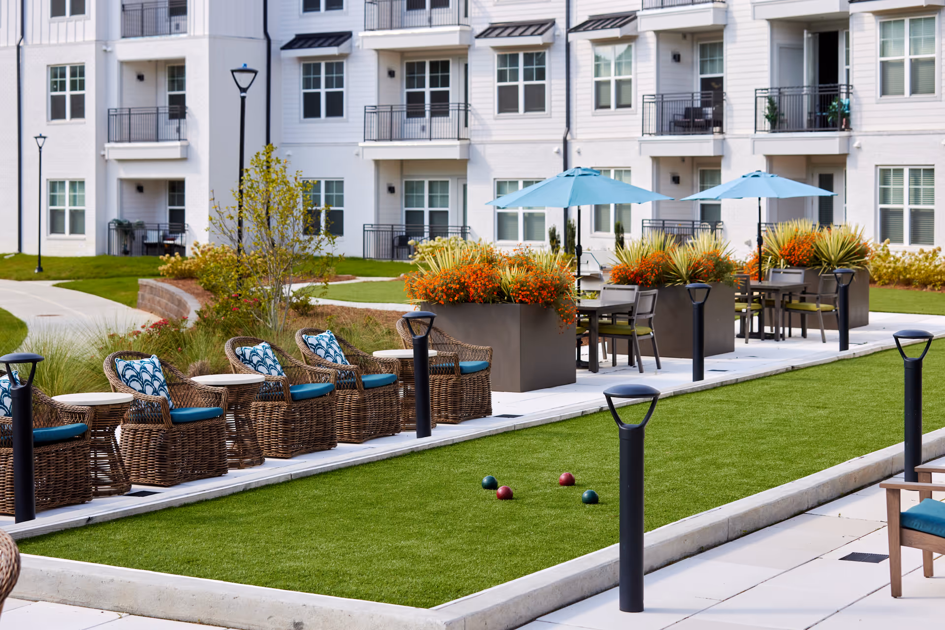 Outdoor seating area at Inspire Royal Park with wicker chairs and blue cushions lined up along a bocce ball court. There are tables with blue umbrellas and large planters filled with orange flowers and greenery. The background shows a multi-story residential building with balconies and windows.
