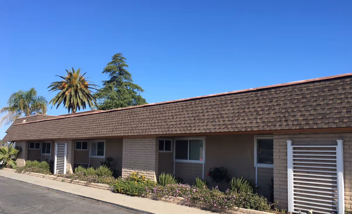 Single-story building facade with a shingled roof, windows, landscaped flowerbeds, and palm trees under a clear blue sky.