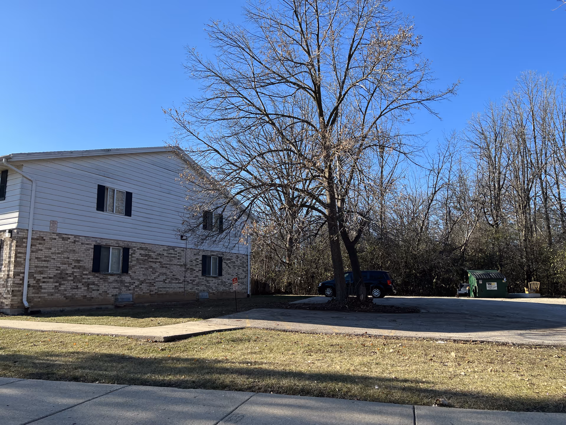 Exterior view of a two-story building with white siding and brick lower walls, a leafless tree in front, a parked black vehicle, a green dumpster, and a clear blue sky.