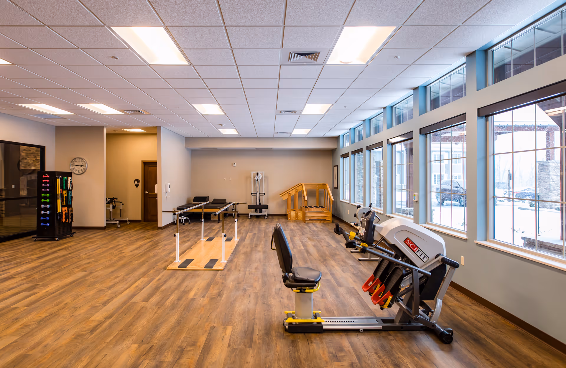 Bright interior therapy/exercise room with large windows, wooden floors, parallel walking bars and fitness equipment.