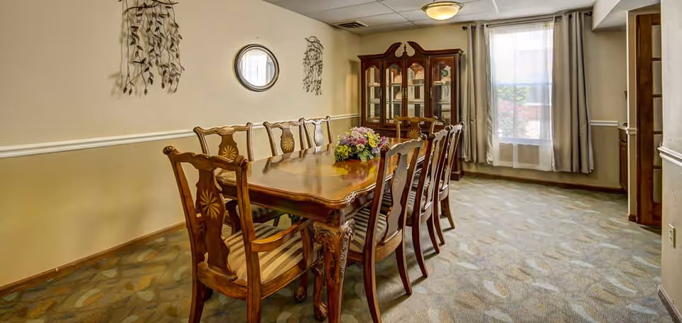 A formal dining room with a long wooden dining table surrounded by eight matching wooden chairs with striped cushions. A floral centerpiece is placed on the table. The room has beige walls with a white chair rail, a patterned carpet, a wooden china cabinet against the far wall, and a window with sheer and gray curtains letting in natural light. Decorative wall hangings and a round mirror are on the left wall.