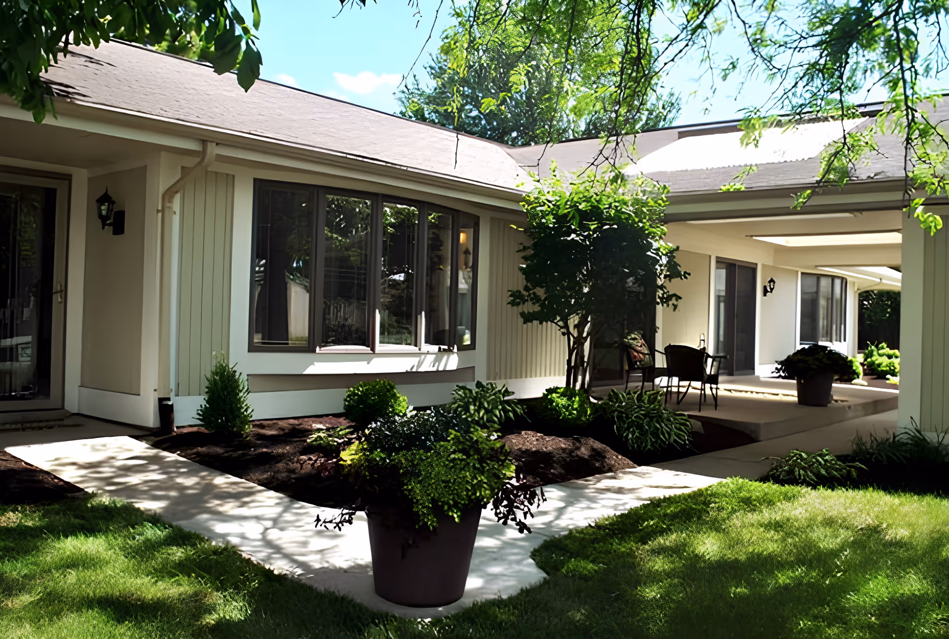 Outdoor view of a senior living facility courtyard at Friendship Village featuring a well-maintained garden with green plants and shrubs, a concrete pathway, and a covered patio area with chairs and a table under a roof extension.