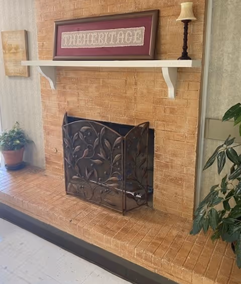 A brick fireplace with a decorative metal screen featuring a leaf and bird design. Above the fireplace is a white mantel shelf holding a framed artwork with the word 'THE HERITAGE' and a small lamp with a beige shade. There are potted plants on either side of the fireplace, one on the floor and one partially visible on the right side.