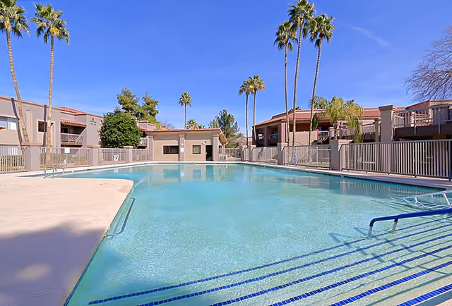 Outdoor swimming pool at Pueblo Norte Senior Living with clear blue water, surrounded by a concrete deck and safety fencing. Palm trees and residential buildings are visible in the background under a clear blue sky.