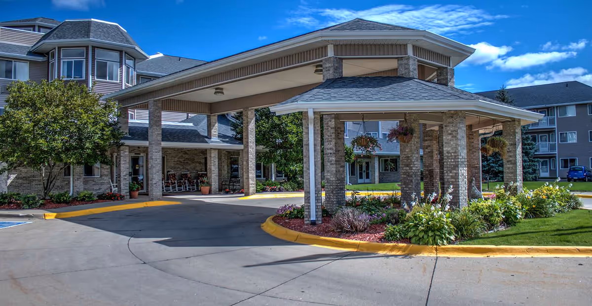 Entrance of a senior living facility with a covered driveway supported by brick pillars, surrounded by landscaped greenery and flowers under a blue sky with some clouds.