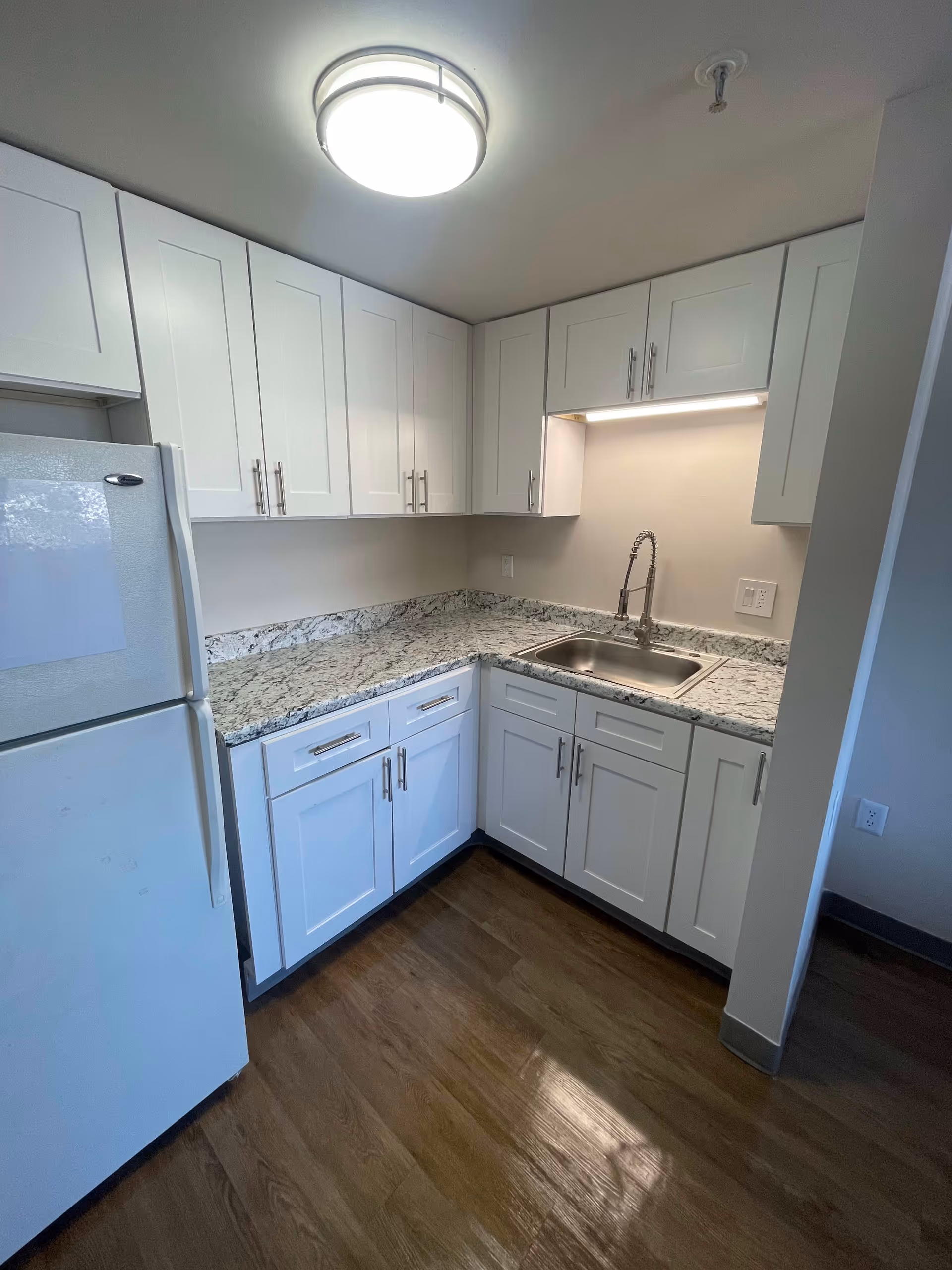 A small kitchen area with white cabinets, a granite countertop, a stainless steel sink with a modern faucet, a white refrigerator, and wood flooring. The kitchen is well-lit with a ceiling light and under-cabinet lighting.