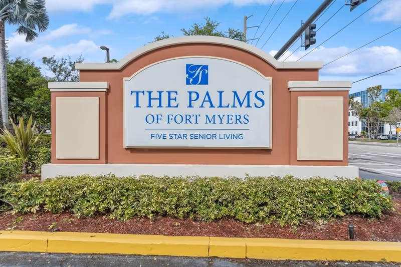 Outdoor sign for The Palms of Fort Myers, a five star senior living facility, surrounded by landscaping with bushes and mulch, located near a street with traffic lights and buildings in the background.