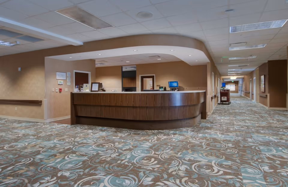 A curved reception desk in a spacious interior with patterned carpet and a long corridor in a senior living facility.