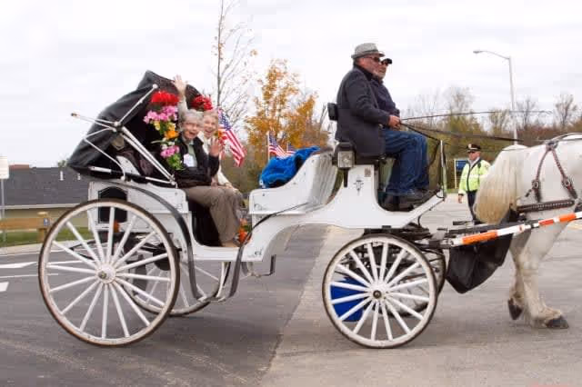 Two elderly women sitting in a white horse-drawn carriage decorated with flowers and small American flags, being driven by a man wearing a hat and sunglasses on a paved road with a police officer standing in the background.