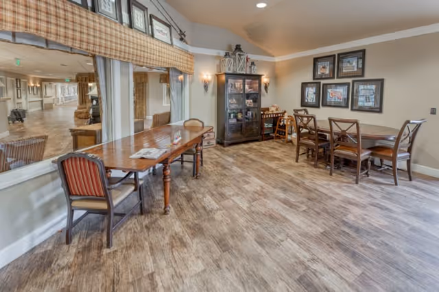 A cozy senior living common area with wooden floors, a long wooden table with chairs, a smaller dining table with four chairs, a glass-front cabinet with decorative items, framed pictures on the wall, and a large window looking into a hallway.