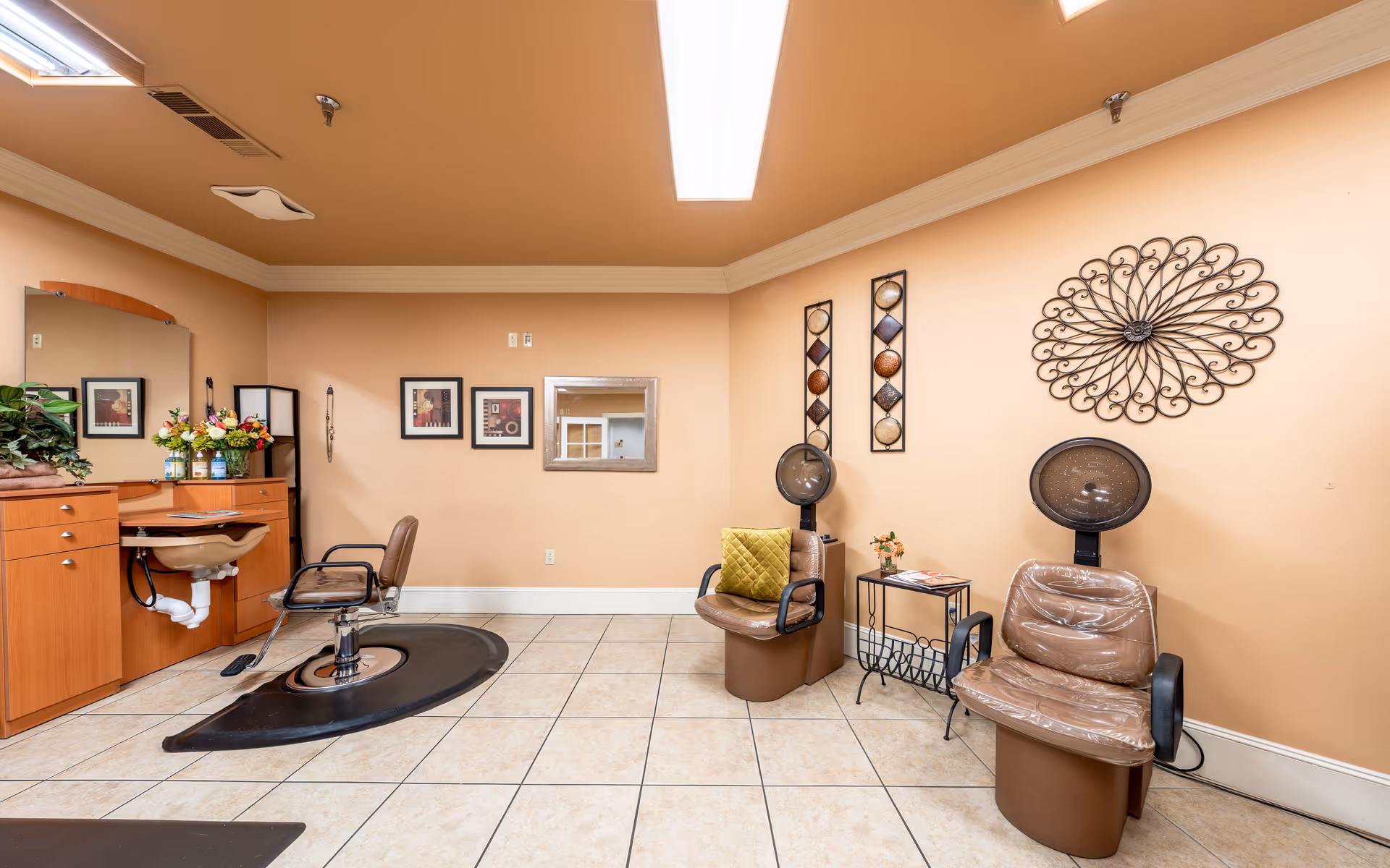 Interior view of a salon area with a hair styling chair in front of a mirror and sink on the left, and two brown salon chairs with hair dryers on the right. The walls are beige with decorative wall art, and the floor is tiled.