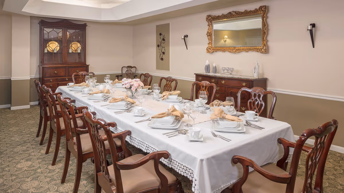 A formal dining room with a long rectangular table covered with a white tablecloth and set with plates, glasses, napkins, and silverware. The table is surrounded by wooden chairs with cushioned seats. In the background, there is a wooden cabinet with glass doors displaying plates, a sideboard with decorative items, and a large ornate gold-framed mirror on the wall.