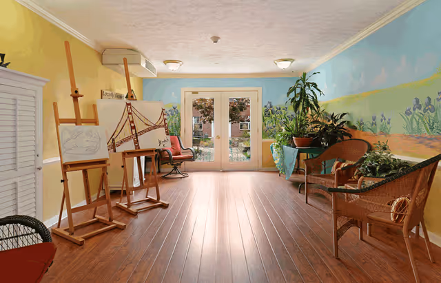 A bright room with wooden flooring featuring three easels holding canvases with artwork, wicker chairs, and a table with potted plants. The walls are painted with a colorful mural depicting a garden scene. Double glass doors at the far end open to an outdoor area with greenery.