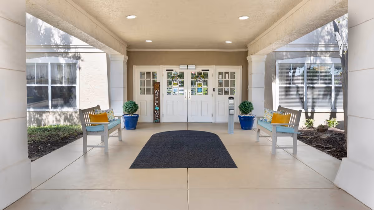 Covered entrance area of a senior living facility with two benches on either side, each with colorful cushions. There are two large potted plants in blue pots flanking double white doors with glass panels. A vertical wooden welcome sign is placed near the left door. The area is well-lit with ceiling lights and has a dark floor mat in front of the doors.